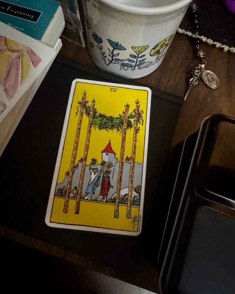 A card lays on an altar. It’s yellow, with four large staffs in the foreground. They are decorated with greenery. There are two people dressed up and look to be celebrating in the middle ground. In the background is a castle, and people heading in that direction. 